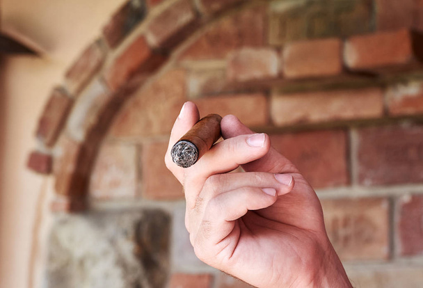 A man holds a burning cuban cigar in his hand and smoking. Smoker in front of defocused brick wall. - Photo, Image