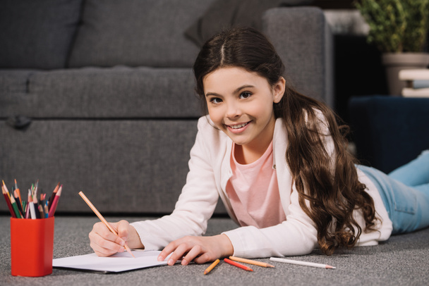 cheerful kid lying on floor and drawing on paper in living room  - Foto, Imagen