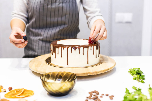 Confectioner decorates with berries a biscuit cake with white cream and chocolate. Cake stands on a wooden stand on a white table. The concept of homemade pastry, cooking cakes. - Foto, Bild