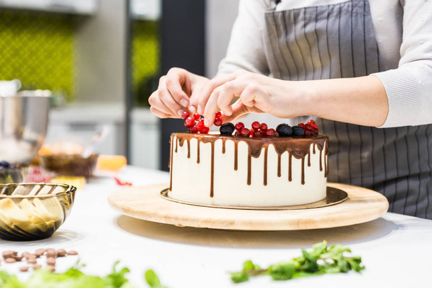Confectioner decorates with berries a biscuit cake with white cream and chocolate. Cake stands on a wooden stand on a white table. The concept of homemade pastry, cooking cakes. - Foto, Imagem