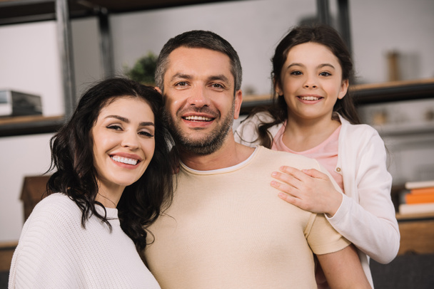 homem feliz sorrindo perto de esposa atraente e filha alegre em casa
  - Foto, Imagem