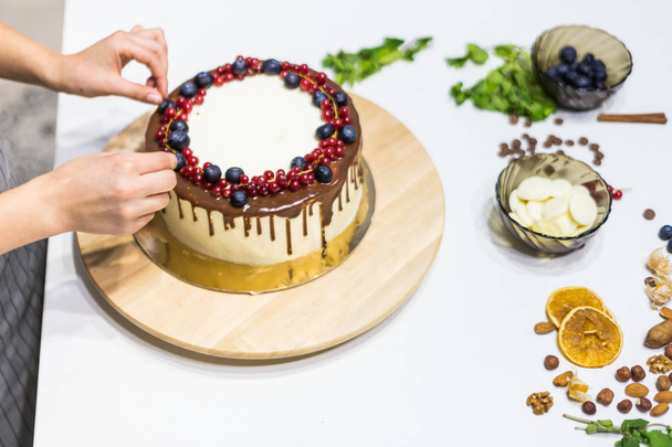 Confectioner decorates with berries a biscuit cake with white cream and chocolate. Cake stands on a wooden stand on a white table. The concept of homemade pastry, cooking cakes. - Φωτογραφία, εικόνα