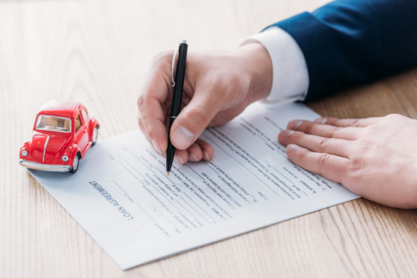 gedeeltelijke weergave autodealer schrijven in leningsovereenkomst in de buurt van rode miniatuur auto op houten tafelblad - Foto, afbeelding