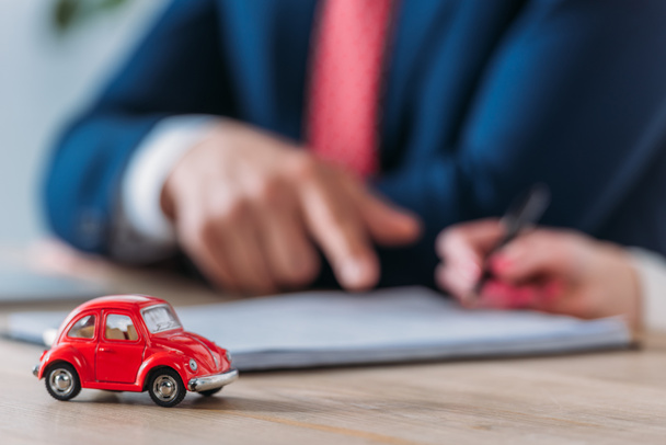 περικομμένη βολή του πελάτη που κρατά στυλό και πωλητή αυτοκινήτων που δείχνει στο σημείο υπογραφής σε σύμβαση κοντά σε κόκκινο αυτοκίνητο παιχνίδι στο τραπέζι - Φωτογραφία, εικόνα