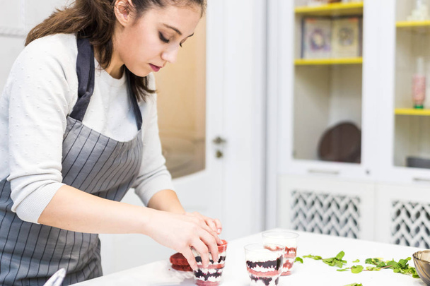 A confectioner prepares a trifle in three cups. Desserts are on the white table in the kitchen. The concept of homemade pastry, cooking cakes. - Photo, Image