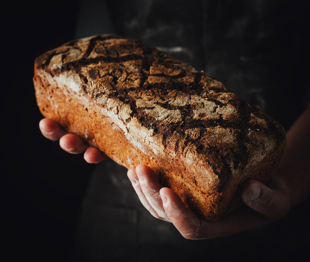 Female hands hold dark rye bread. Baker with fresh bread on a dark background - Photo, Image