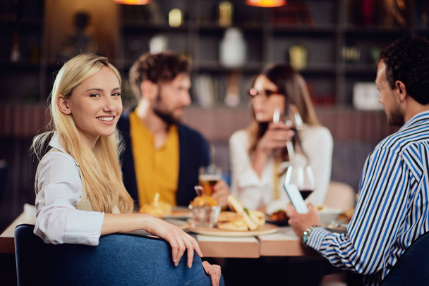 Jonge glimlachende blanke blonde leuke vrouw gekleed slimme casual zitten aan tafel in restaurant en kijken over de schouder. Op de achtergrond zijn haar vrienden aan het eten.. - Foto, afbeelding