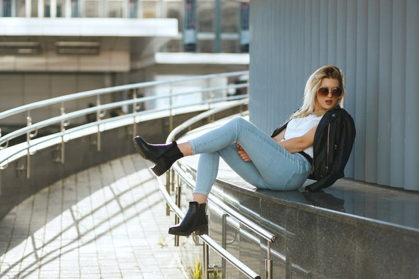 通りに座ってジーンズとジャケットを着てグラマースタイリッシュな女性  - 写真・画像