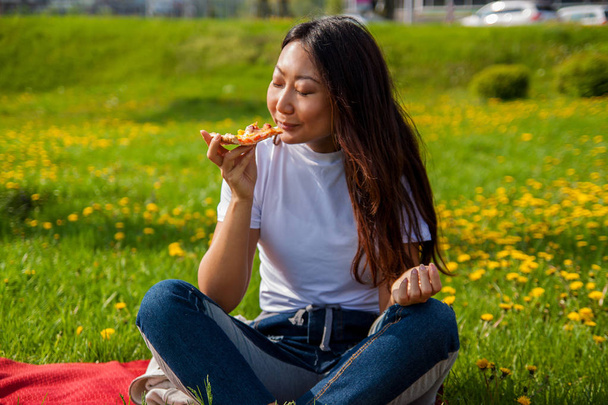 junge asiatische Frau mit langen Haaren, die ein Stück Pizza hält, während sie auf dem Gras sitzt und das Mittagessen genießt. - Foto, Bild