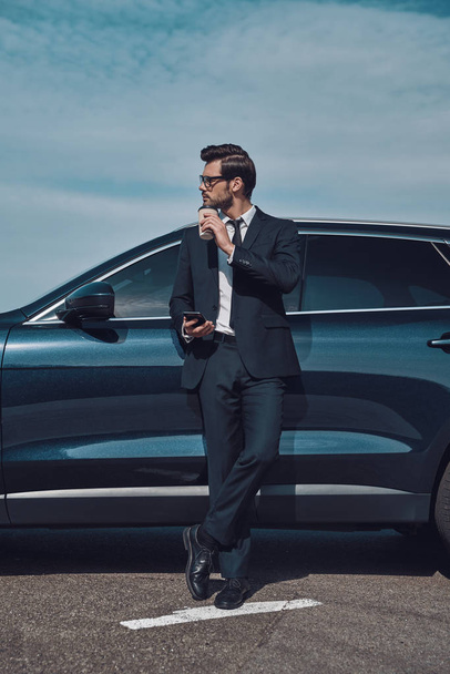 Time for a little break. Full length of handsome young businessman using smart phone and drinking coffee while standing near his car outdoors - Photo, Image