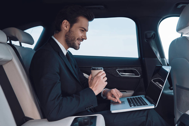 Driven by success. Handsome young man in full suit working using laptop while sitting in the car - Zdjęcie, obraz