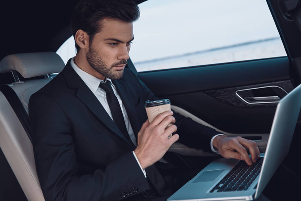 Working from car. Handsome young man in full suit working using laptop while sitting in the car - Foto, afbeelding