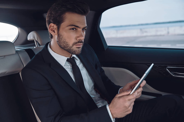 Full concentration at work. Handsome young man in full suit working using digital tablet while sitting in the car           - Foto, afbeelding