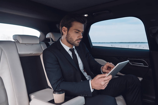 Considering the next step. Handsome young man in full suit working using digital tablet while sitting in the car     - Fotografie, Obrázek