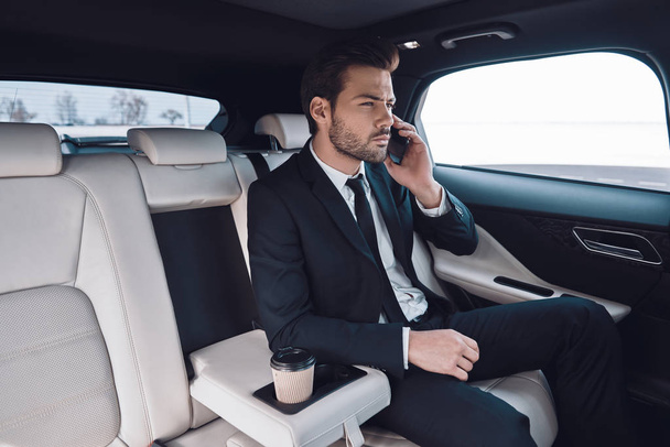 Business on the move. Handsome young man in full suit talking on the smart phone while sitting in the car - Foto, Imagem