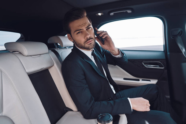 Busy day. Handsome young man in full suit talking on the smart phone and looking at camera while sitting in the car - Foto, Imagem