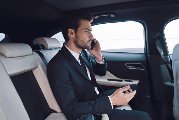 Always available. Handsome young man in full suit talking on the smart phone while sitting in the car - Foto, afbeelding