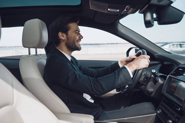 In love with high speed. Handsome young man in full suit smiling while driving a car    - Foto, Bild