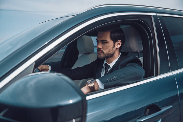 Just keep moving! Handsome young man in full suit looking straight while driving a car                      - Фото, зображення