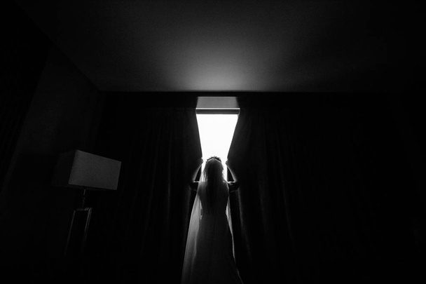 πίσω όψη της νεαρής νύφης στο νυφικό και πέπλο άνοιγμα κουρτίνες, μαύρο και άσπρο εικόνα - Φωτογραφία, εικόνα