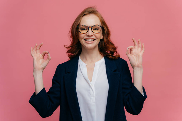 Pleased female financier makes okay gesture with both hands, has pleasant smile on face, wears formal wear, has positive smile, models over pink background, expresses satisfaction or agreement - Photo, Image