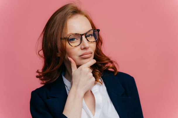 Thoughtful red haired lady holds chin, looks with contemplative expression at camera, thinks over future plans, has full lips, healthy skin, dressed in formal apparel, stands against pink background - Foto, Imagem
