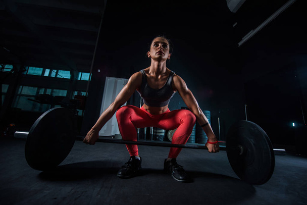 Young, European, muscular girl in red leggings, doing exercise with a barbell in the gym for crossfit. Dark background - 写真・画像