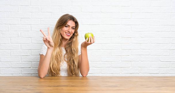 Mladý blondýnka žena s jablko dělat vítězství gesto - Fotografie, Obrázek