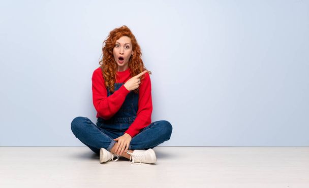 Redhead woman with overalls sitting on the floor surprised and pointing side - Photo, Image