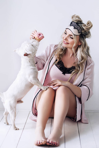 Beautiful european woman with her dog in white apartment, european woman is playing with her white dog, pretty woman with light hair in silk pijamas in white apartment, beautiful pretty woman with - Foto, imagen