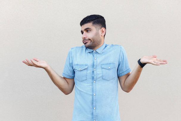 confused handsome young bearded man in blue shirt standing with raised arms and looking aside on light beige wall background. - Foto, afbeelding