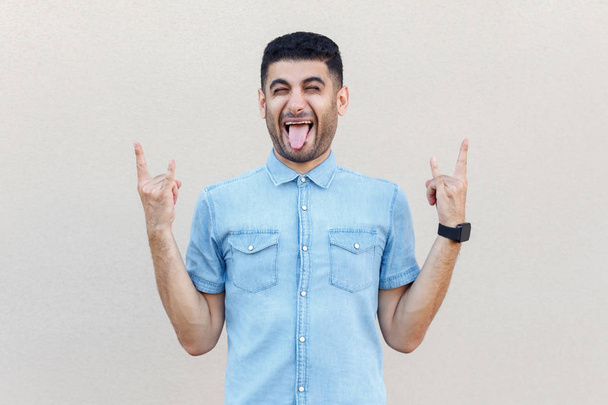  funny crazy handsome young bearded man in blue shirt showing rock signs and tongue out on light beige wall background. - 写真・画像