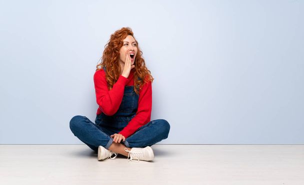Redhead woman with overalls sitting on the floor shouting with mouth wide open to the lateral - Фото, изображение