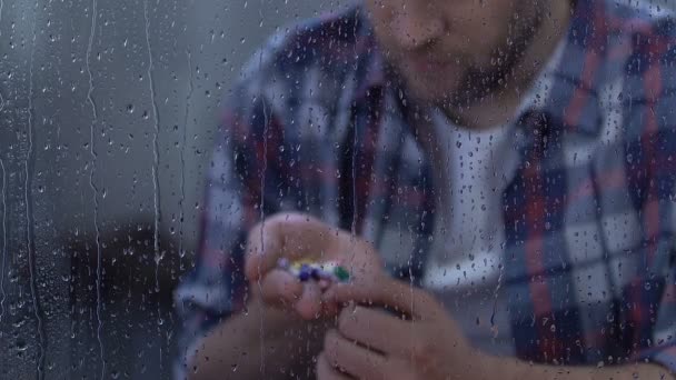 Depressed sick man holding full palm of medication, sitting behind rainy window - Filmati, video