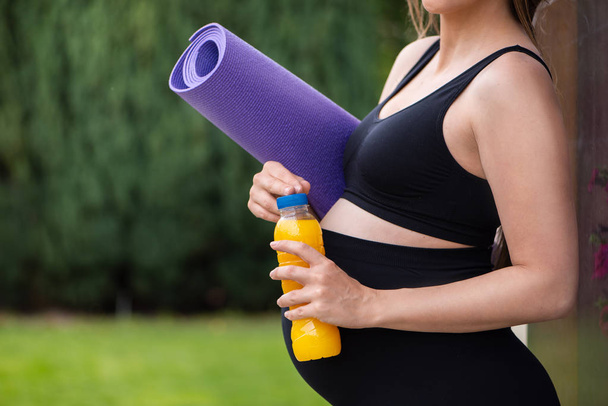 Terhes sportos nő egy lila szőnyeget a jóga a kezében, álló udvarban, ivás narancslé. - Fotó, kép