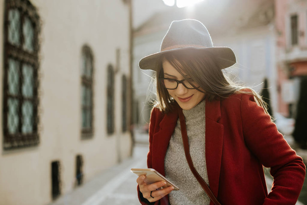 Jovem hipster mulher com óculos segurando um smartphone e usando um chapéu, na cidade velha, Europa
.  - Foto, Imagem