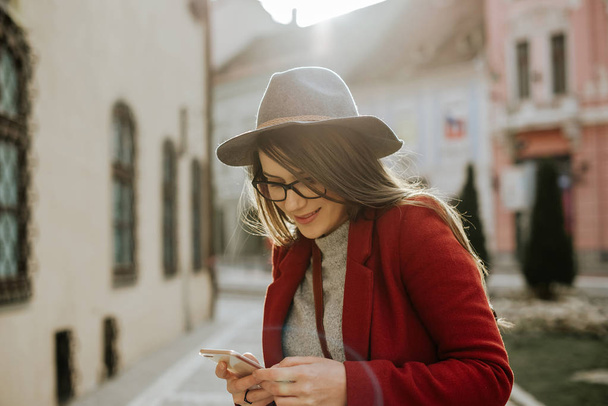Young hipster woman with eyeglasses holding a smartphone and wearing a hat, looking side in the city old town, Europe.  - Photo, Image