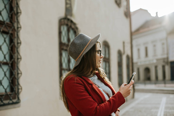 Молодая хипстерша в очках с смартфоном и шляпой, смотрящая в сторону старого города, Европа
.  - Фото, изображение