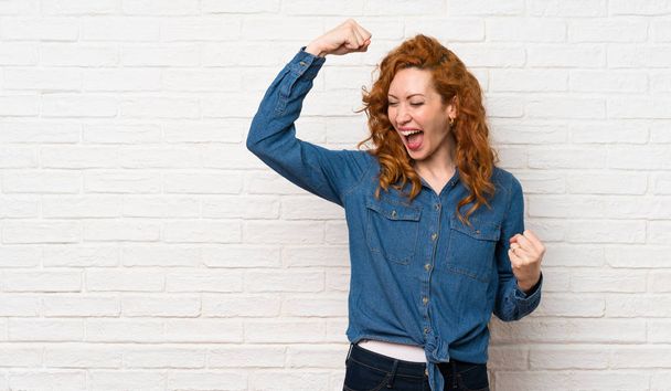 Redhead woman over white brick wall celebrating a victory - Фото, изображение