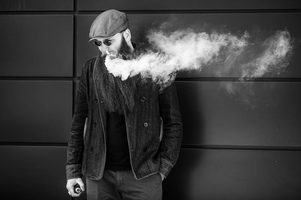 Vape bearded man. An adult man with a very long beard in a cap and sunglasses smokes an electronic cigarette on a sunny day outside. Bad habit that is harmful to health. Black and white. - Fotoğraf, Görsel