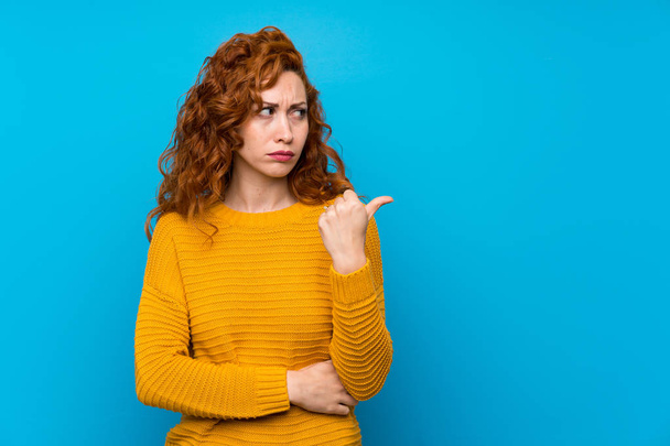 Redhead woman with yellow sweater unhappy and pointing to the side - Photo, Image