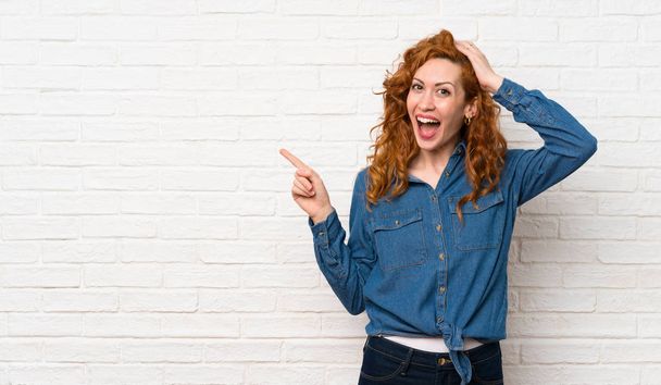 Redhead woman over white brick wall surprised and pointing finger to the side - Foto, Bild