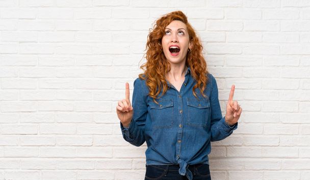 Redhead woman over white brick wall surprised and pointing up - Photo, Image