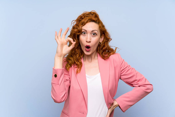 Redhead woman in suit over isolated blue wall surprised and showing ok sign - Photo, Image