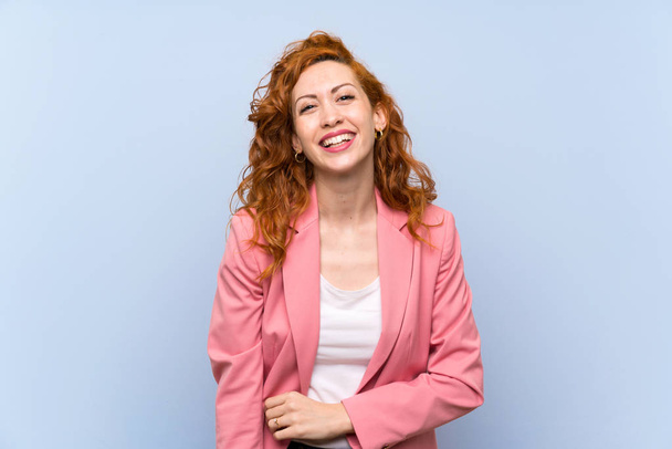 Redhead vrouw in pak over geïsoleerde blauwe muur glimlachend - Foto, afbeelding