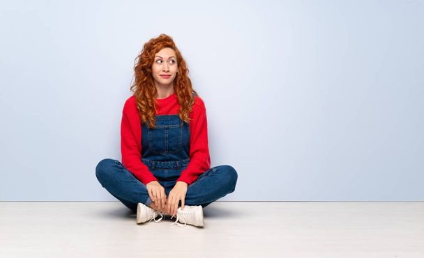 Rudowłosy kobieta z kombinezonu siedzi na podłodze podejmowania wątpliwości gest patrząc strony - Zdjęcie, obraz