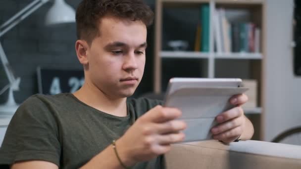 Young teenage boy is concentrated on his tablet, close-up front view in the room - Imágenes, Vídeo