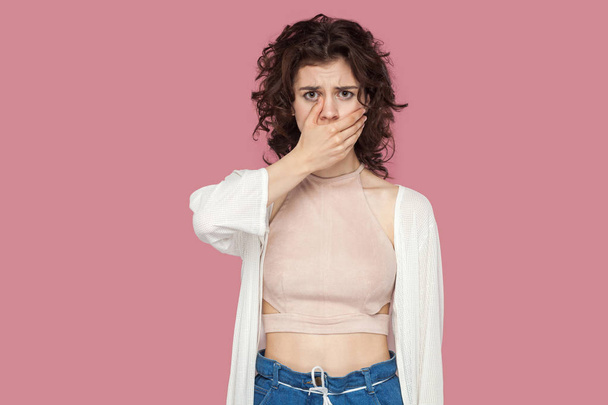 shocked beautiful brunette young woman with curly hairstyle in casual style covering her mouth and looking at camera on pink background. - Zdjęcie, obraz