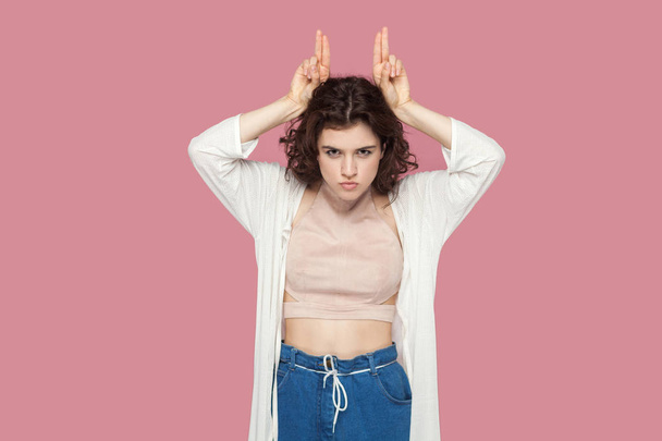 serious funny cute brunette young woman with curly hairstyle in casual style standing with finger horns on her head and looking at camera on pink background. - Photo, image