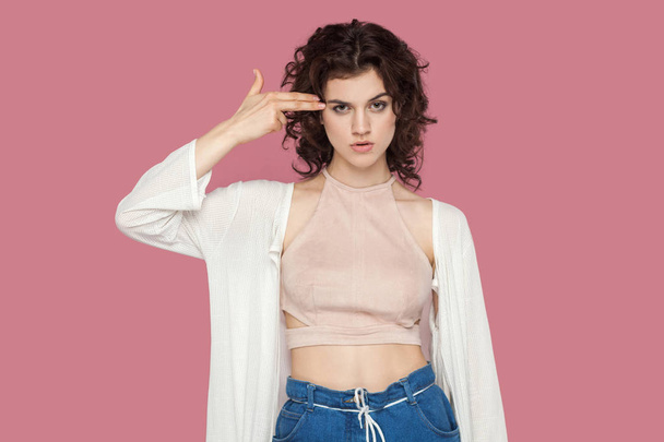 crazy beautiful brunette young woman with curly hairstyle in casual style  standing with pistol gun gesture on head and looking at camera on pink background. - Zdjęcie, obraz